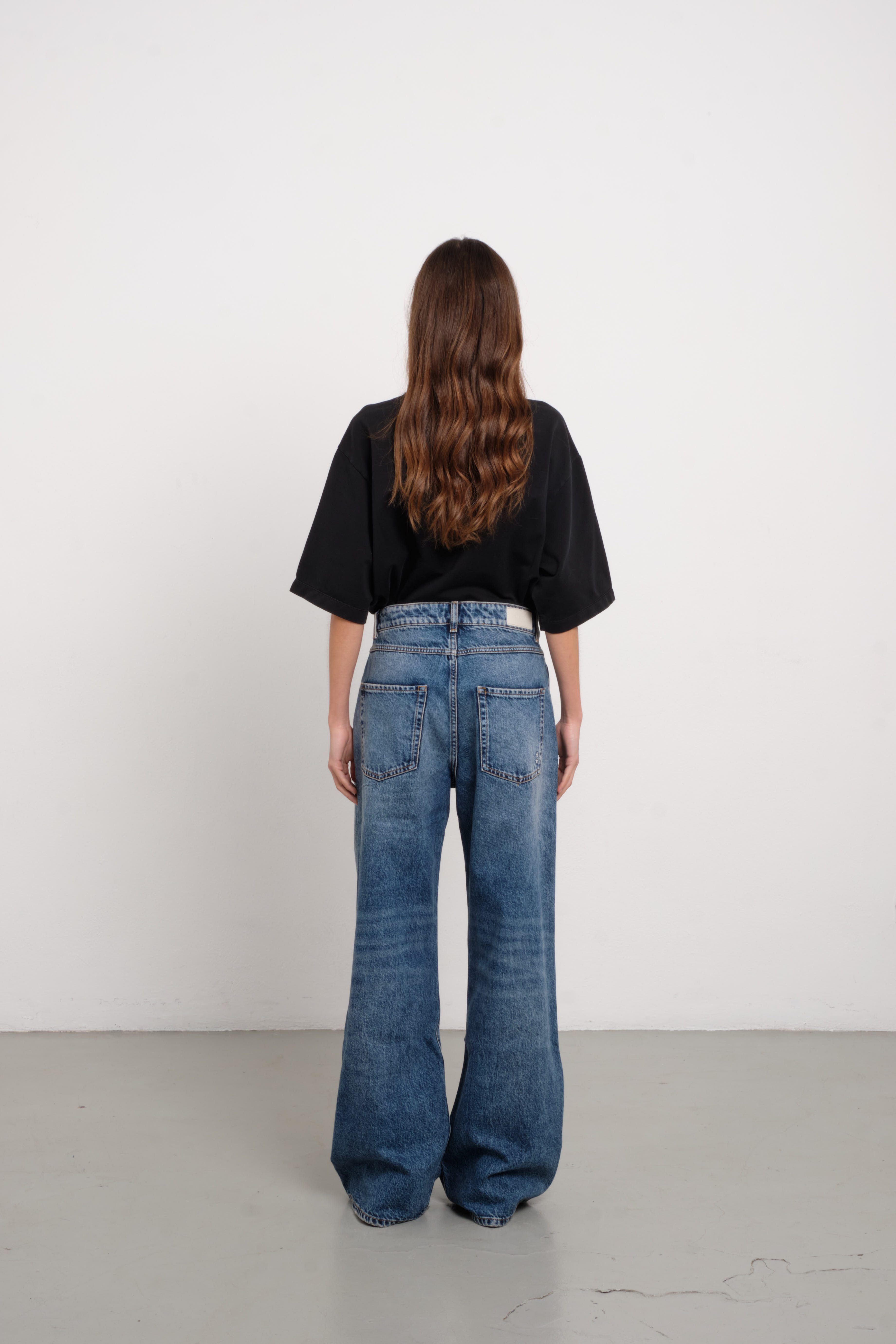 Back view of Debby women’s wide leg jeans in medium wash, 100% rigid cotton denim, worn by a model with a black shirt.