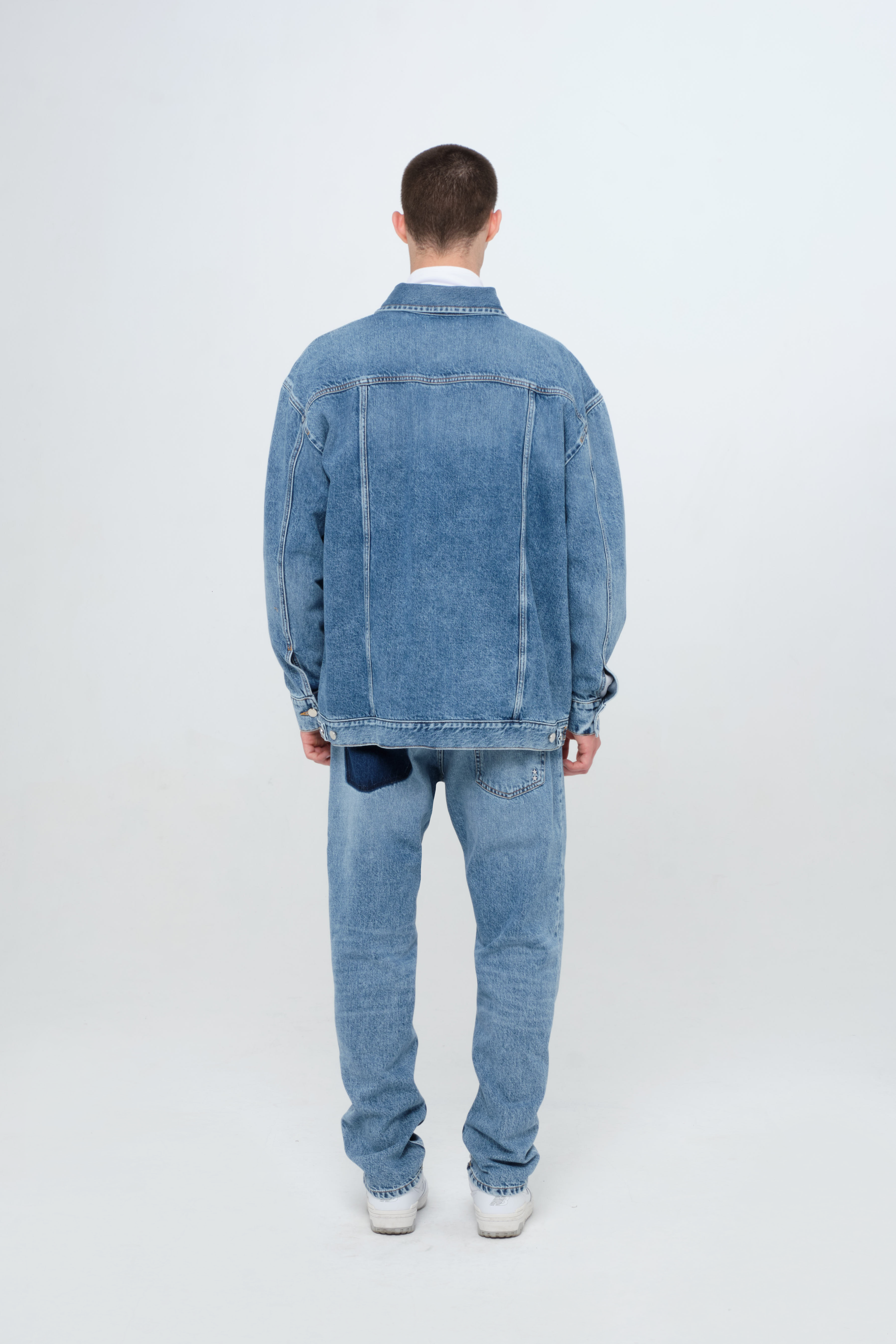 Back view of Drew men’s oversize jacket in medium wash, 100% rigid cotton denim, worn by a model with jeans.