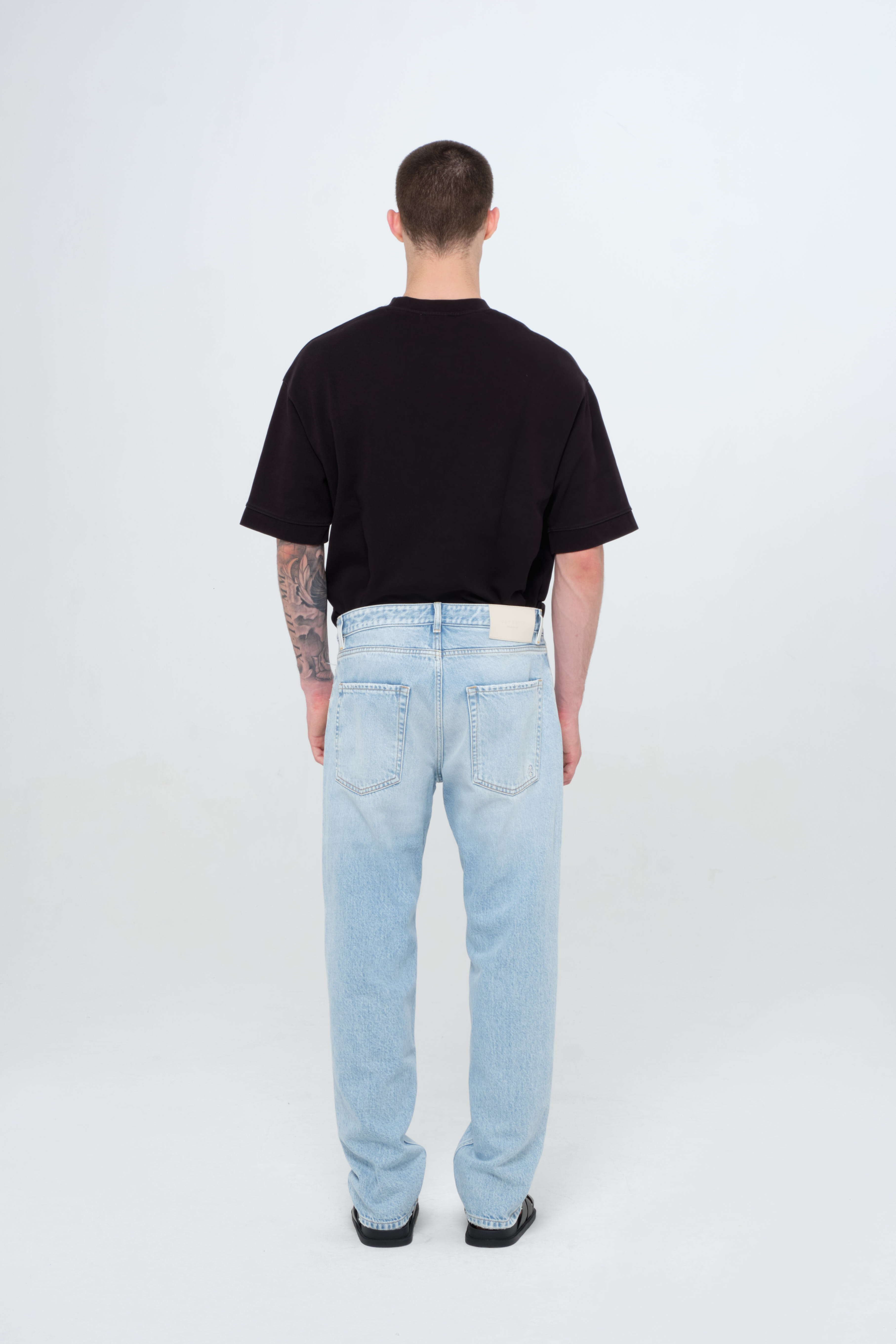 Back view of Kanye men’s loose jeans in light wash, 100% rigid cotton denim, worn by a model with a black t-shirt.