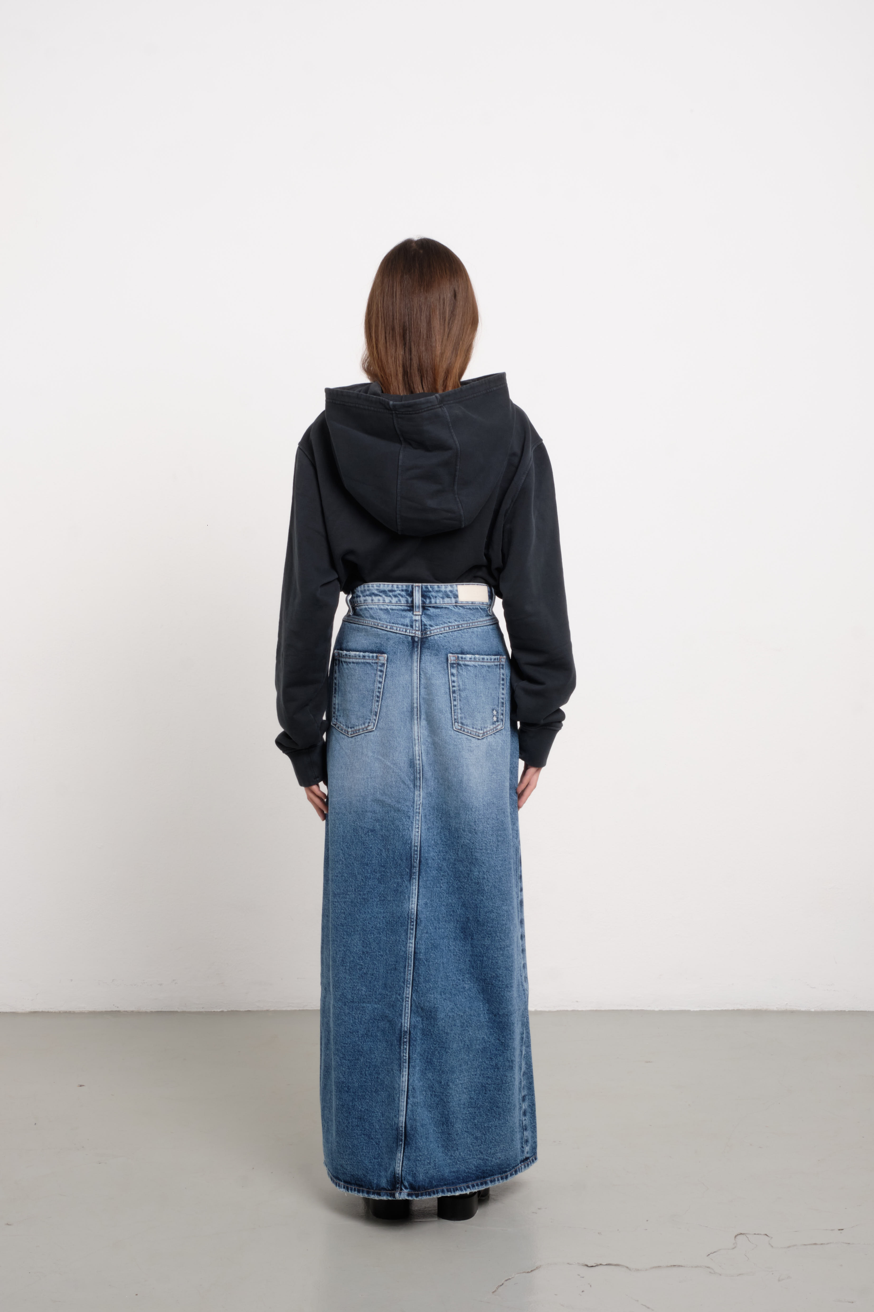 Back view of Lara women’s long skirt in medium wash, 100% rigid cotton denim, worn by a model with a black hoodie.
