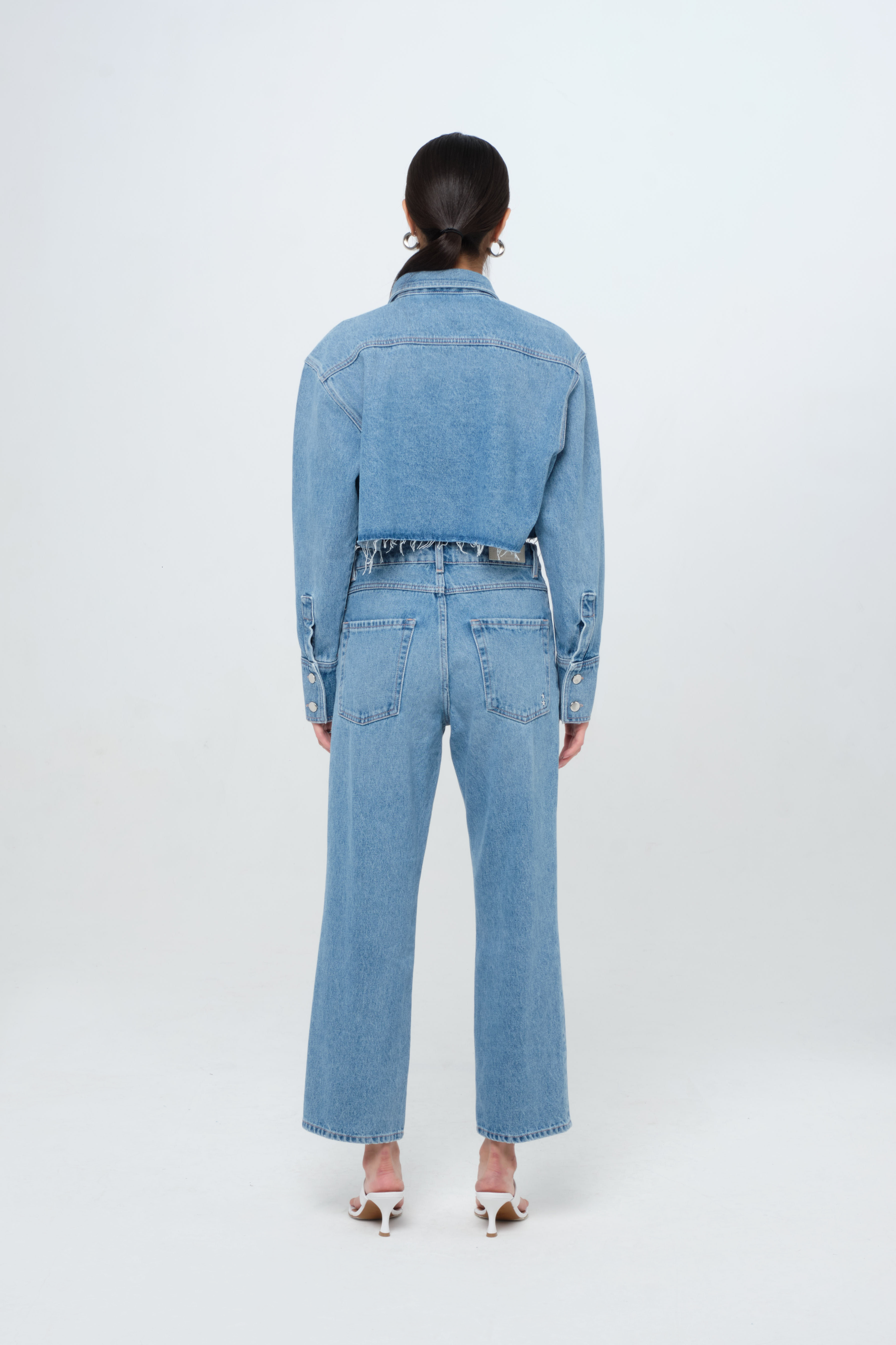 Back view of Amber Eco women’s denim crop shirt in medium wash, 100% rigid organic cotton denim, worn by a model with jeans.
