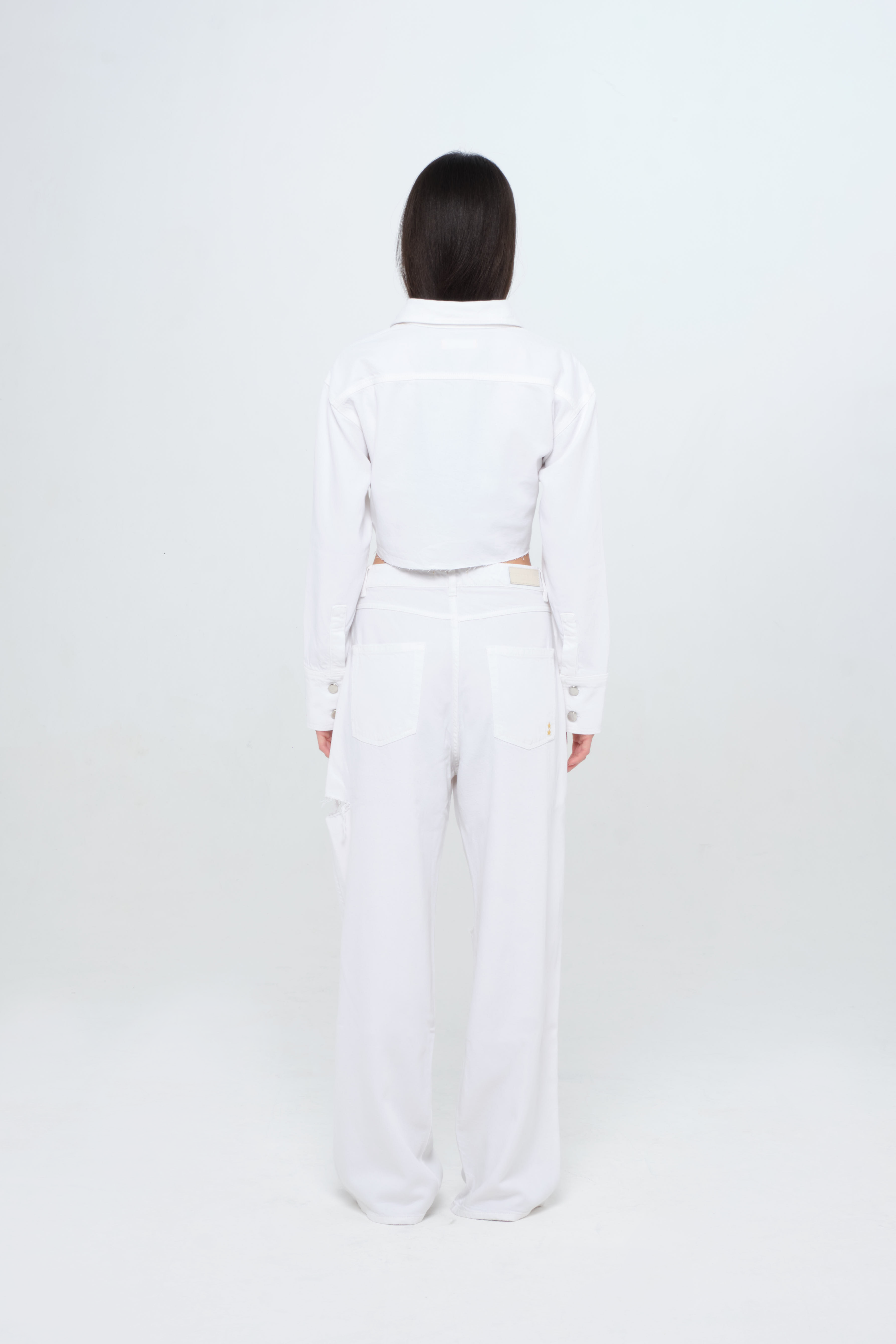 Back view of Amber women’s denim crop shirt in white, 100% rigid soft cotton denim, worn by a model with a white jeans.