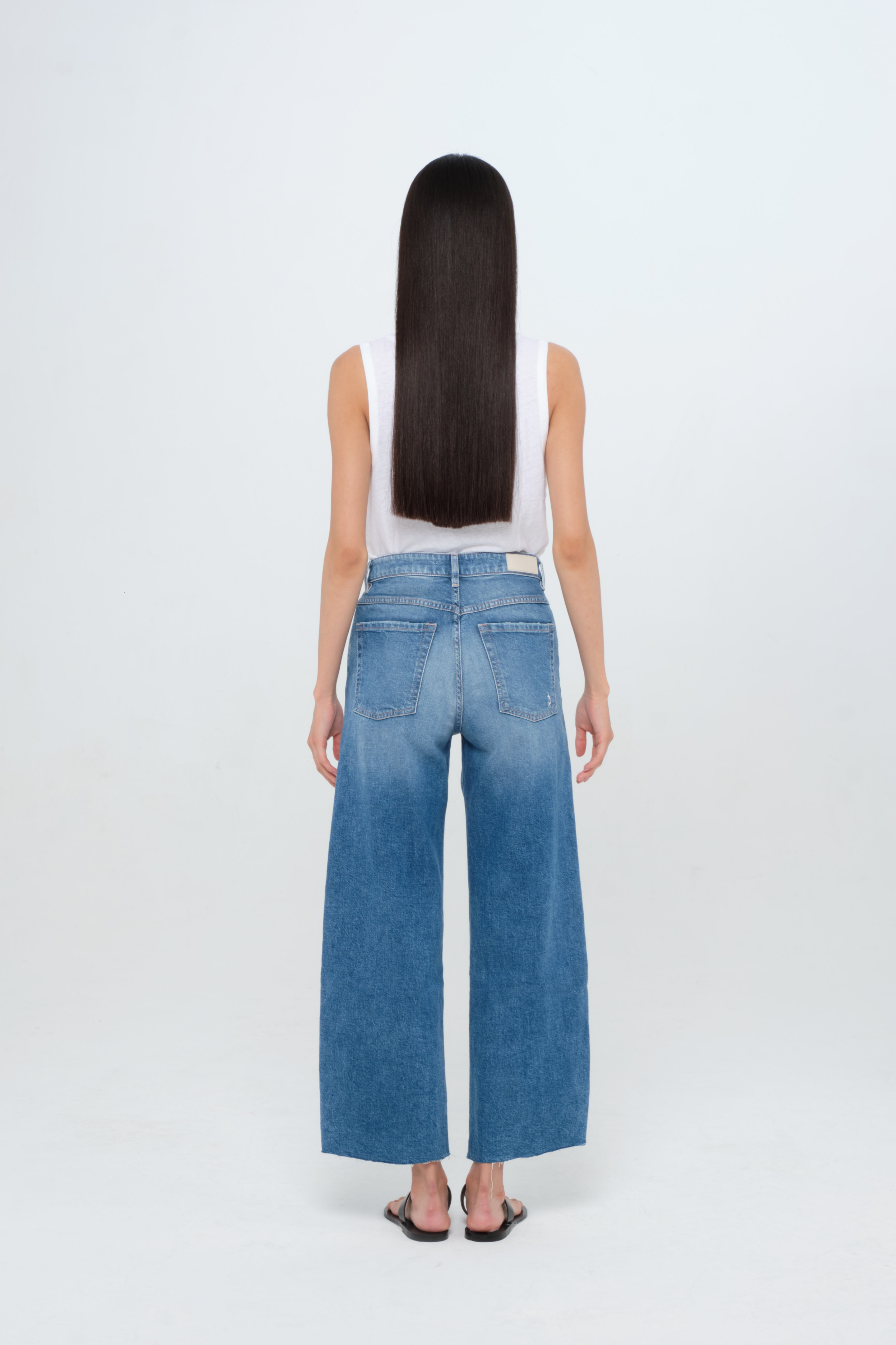 Back view of Angie women’s flared crop jeans in medium wash 100% cotton denim, worn by a model with a white tank top.