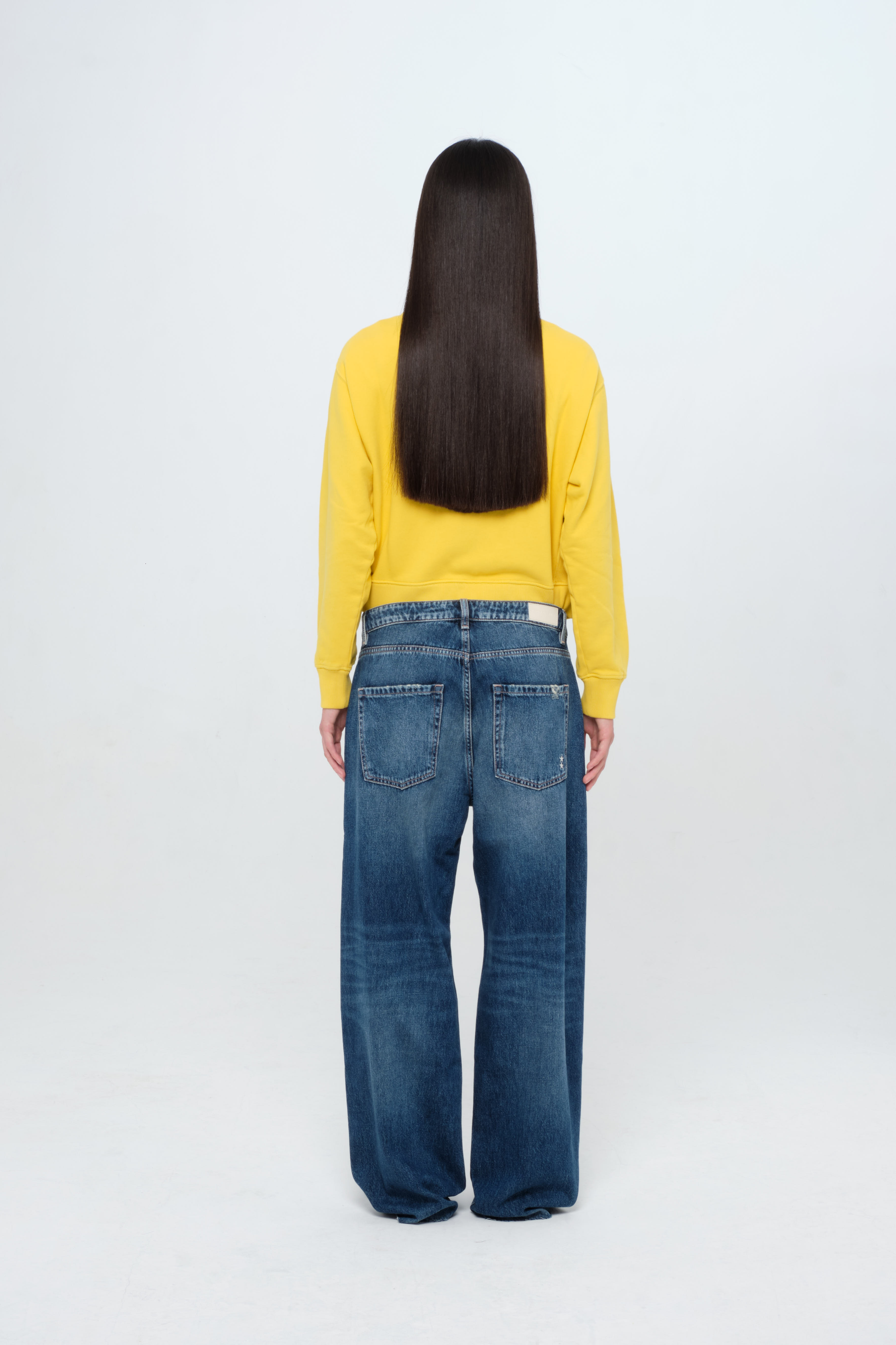 Back view of Bea women’s wide leg jeans in dark wash, 100% rigid cotton denim, worn by a model with a yellow sweatshirt.