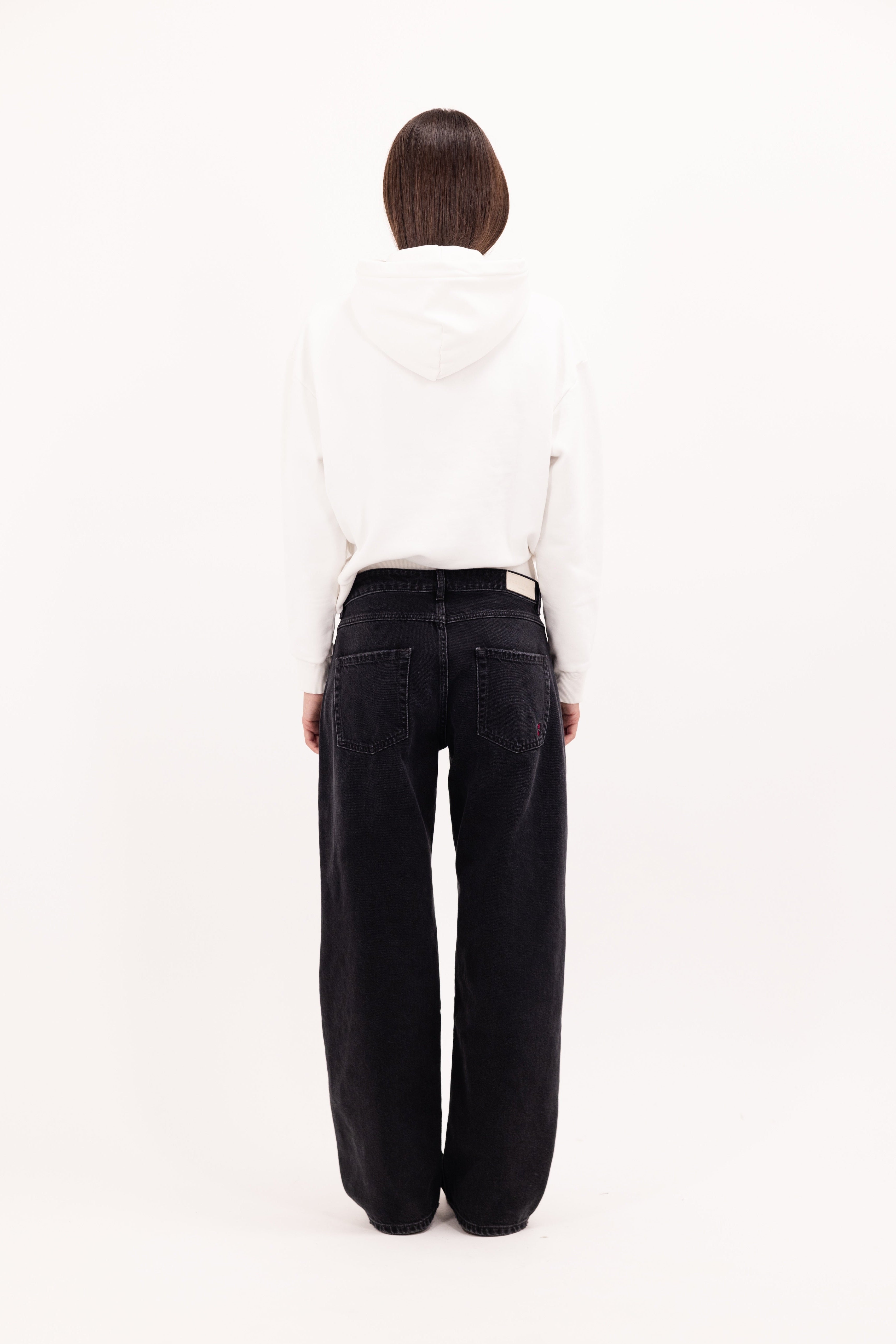 Back view of Bea women’s wide leg jeans in graphite black wash, 100% rigid cotton denim, worn by a model with a white hoodie.