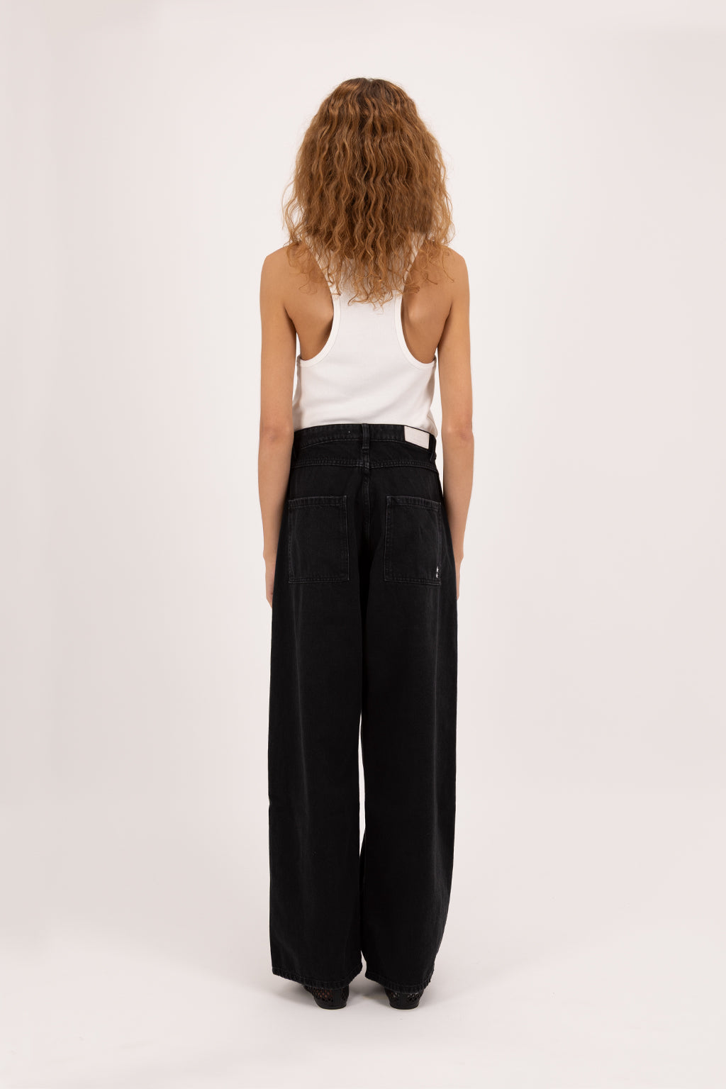 Back view of Bess women’s wide leg jeans in graphite black wash, 100% rigid soft cotton denim, worn by a model with a white tank top.
