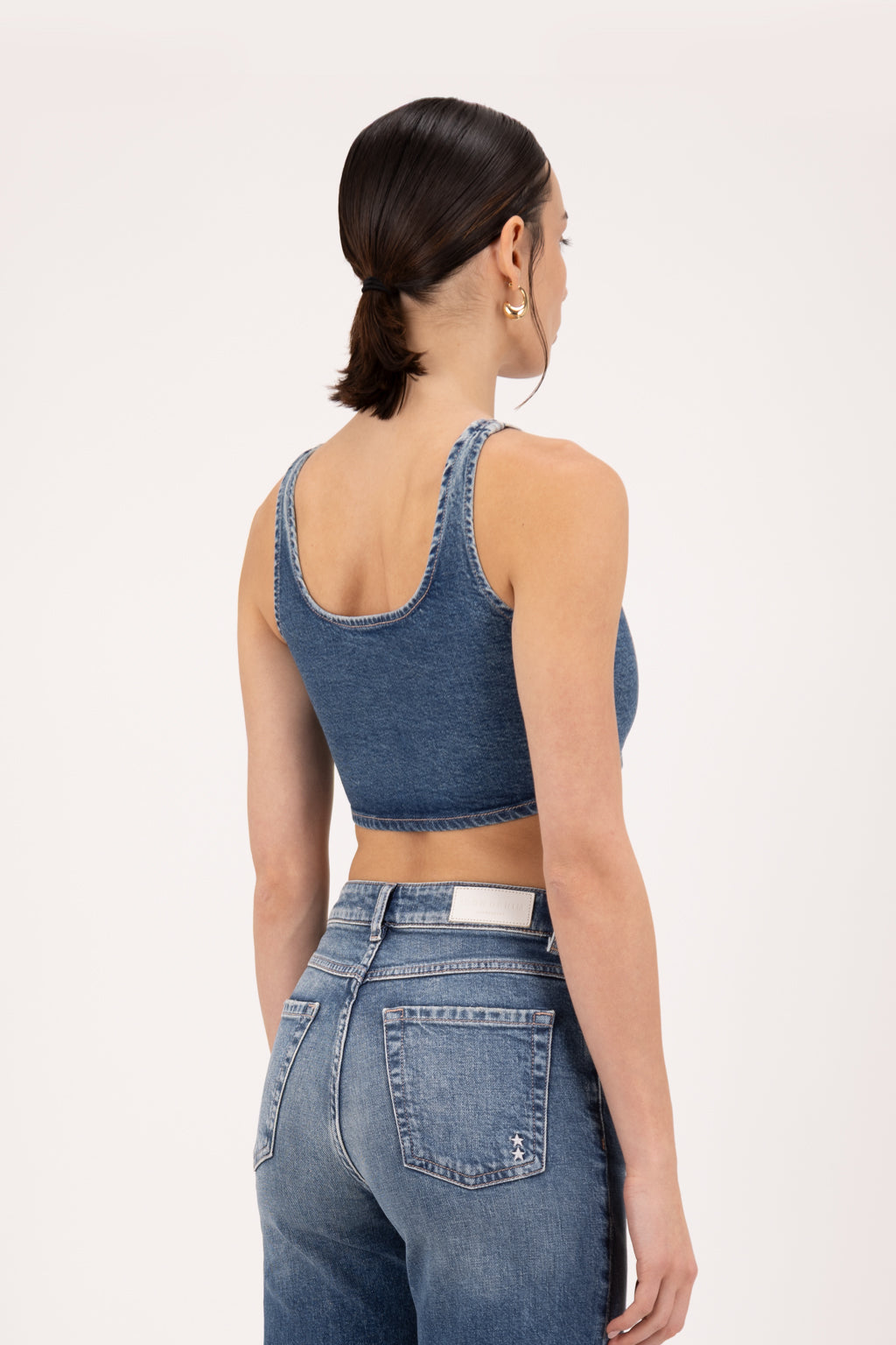 Back view of Bibi women’s crop top in dark used wash, 100% rigid cotton denim, worn by a model with jeans.