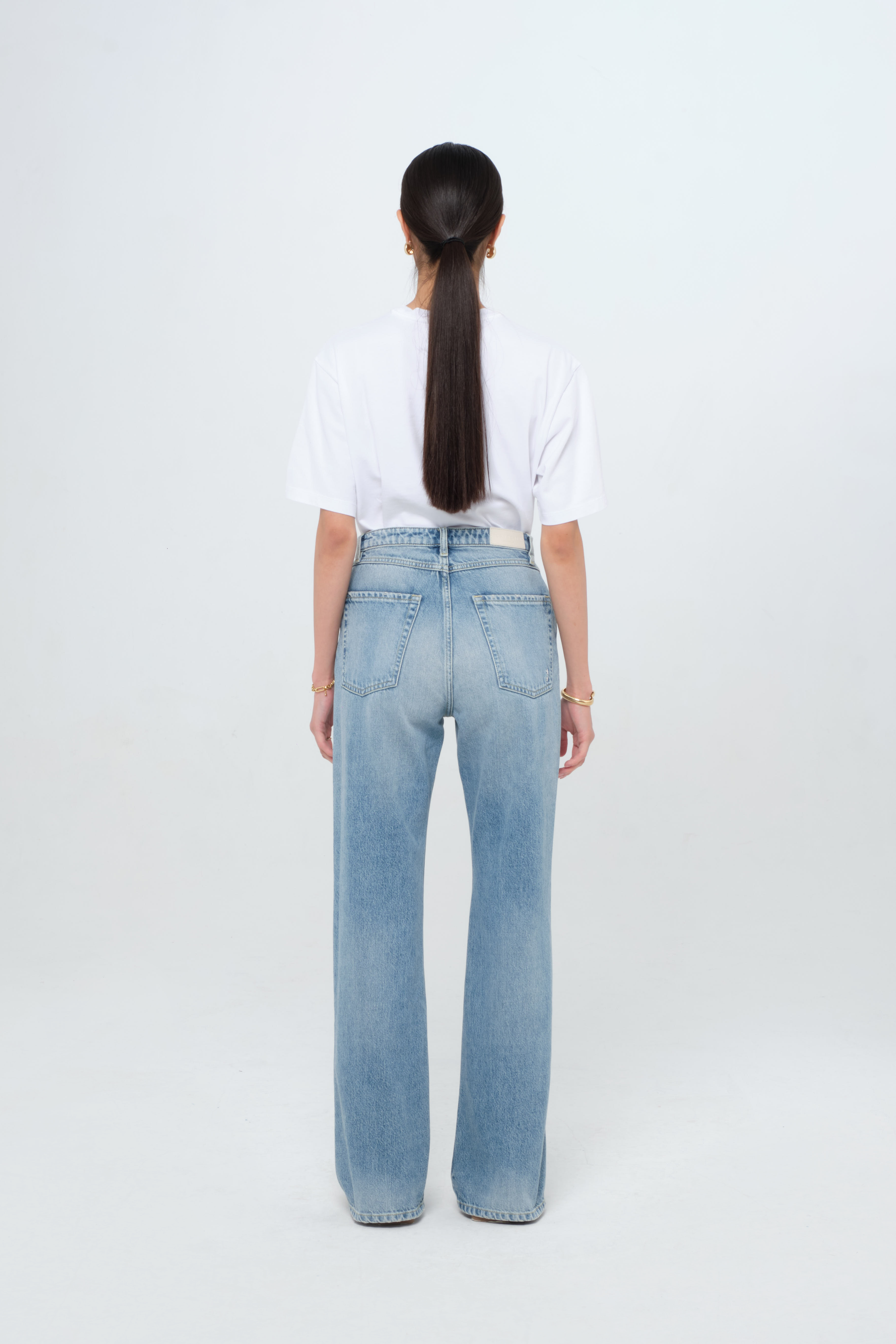 Back view of Blue women’s flared leg jeans in smoke blue wash, 100% rigid cotton denim, worn by a model with a white t-shirt.
