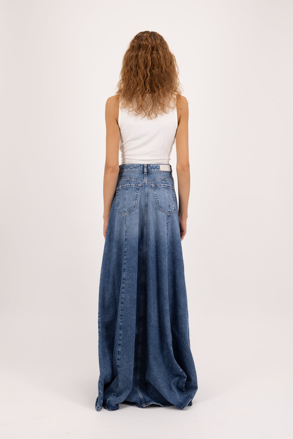 Back view of Cora women’s oversize skirt in eclipse wash, 100% rigid cotton denim, worn by a model with a white tank top.