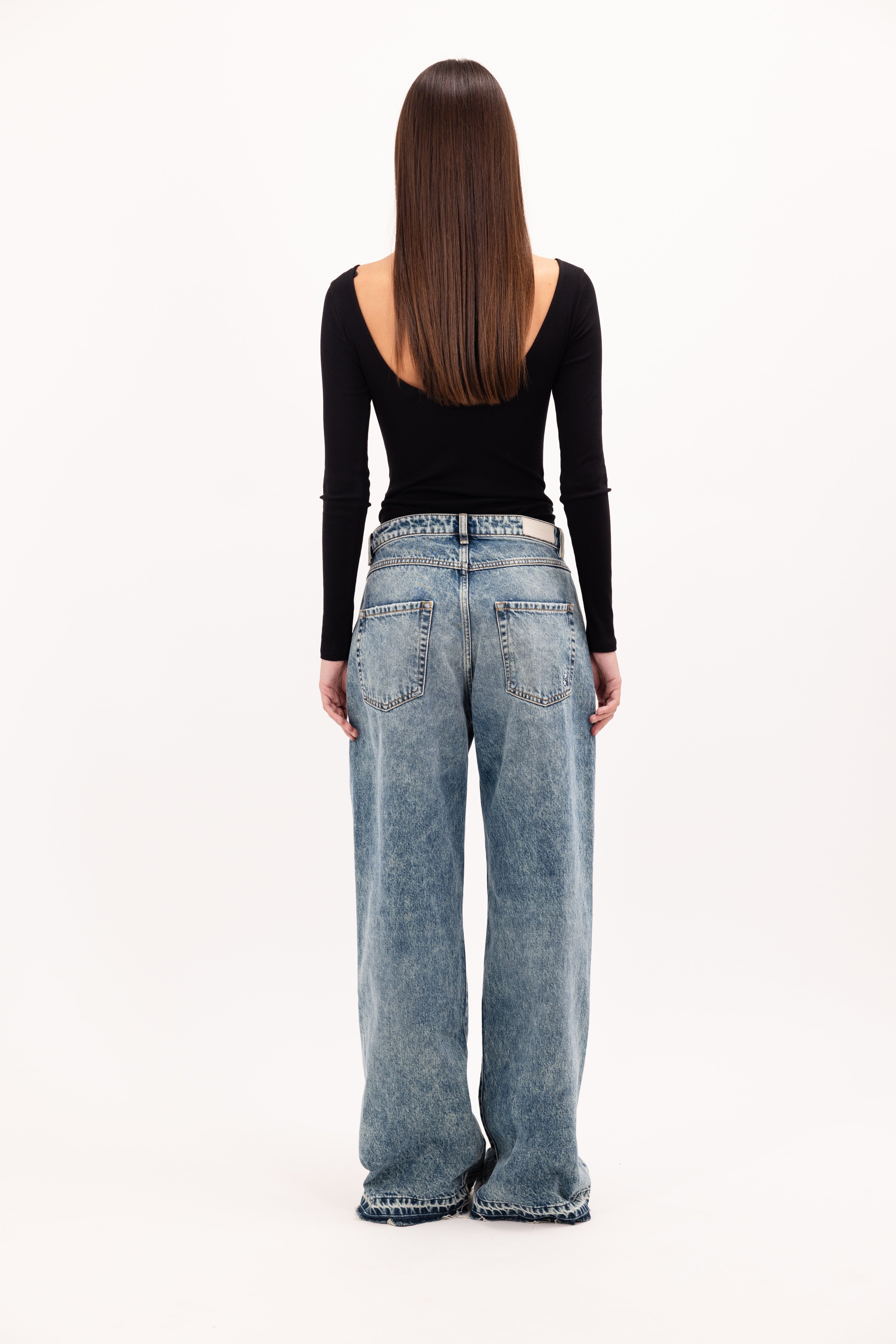 Back view of Debby women’s wide leg jeans in aged stone wash, 100% rigid cotton denim, worn by a model with a black top.