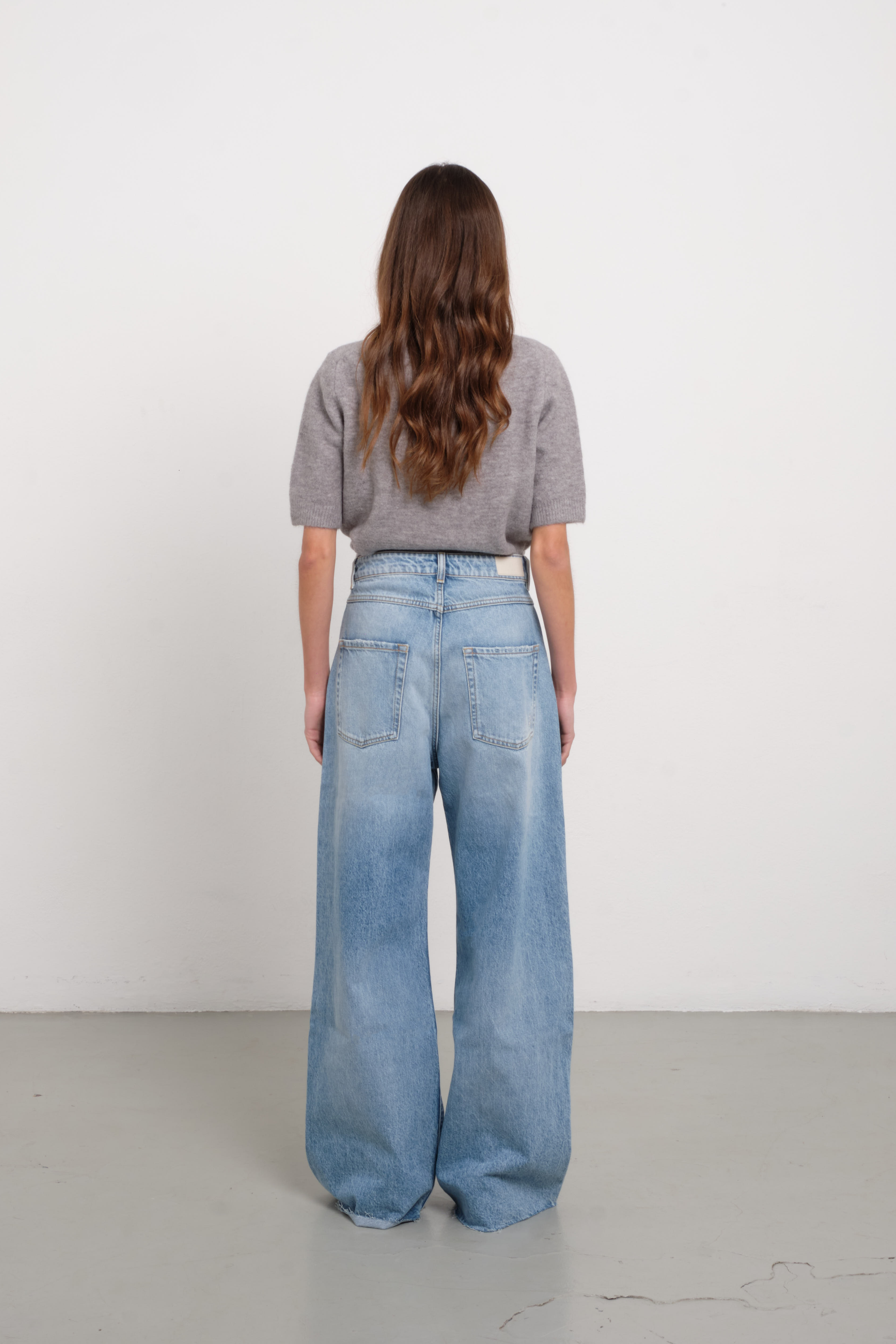 Back view of Debby women’s wide leg jeans in light wash, 100% rigid cotton denim, worn by a model with a gray shirt.
