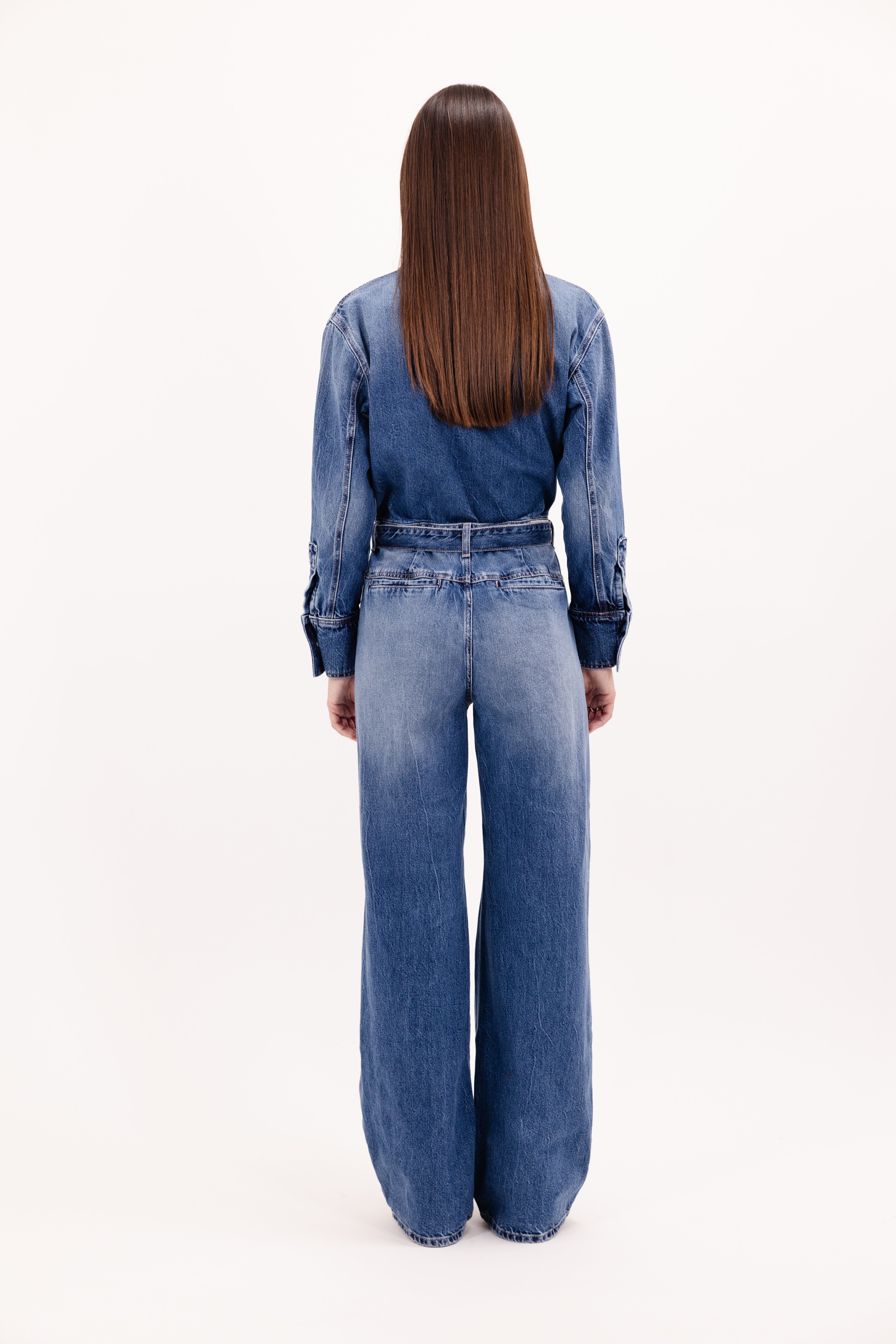 Back view of Indie women’s jumpsuit in eclipse wash, 100% rigid cotton denim, worn by a model with a black pullover.