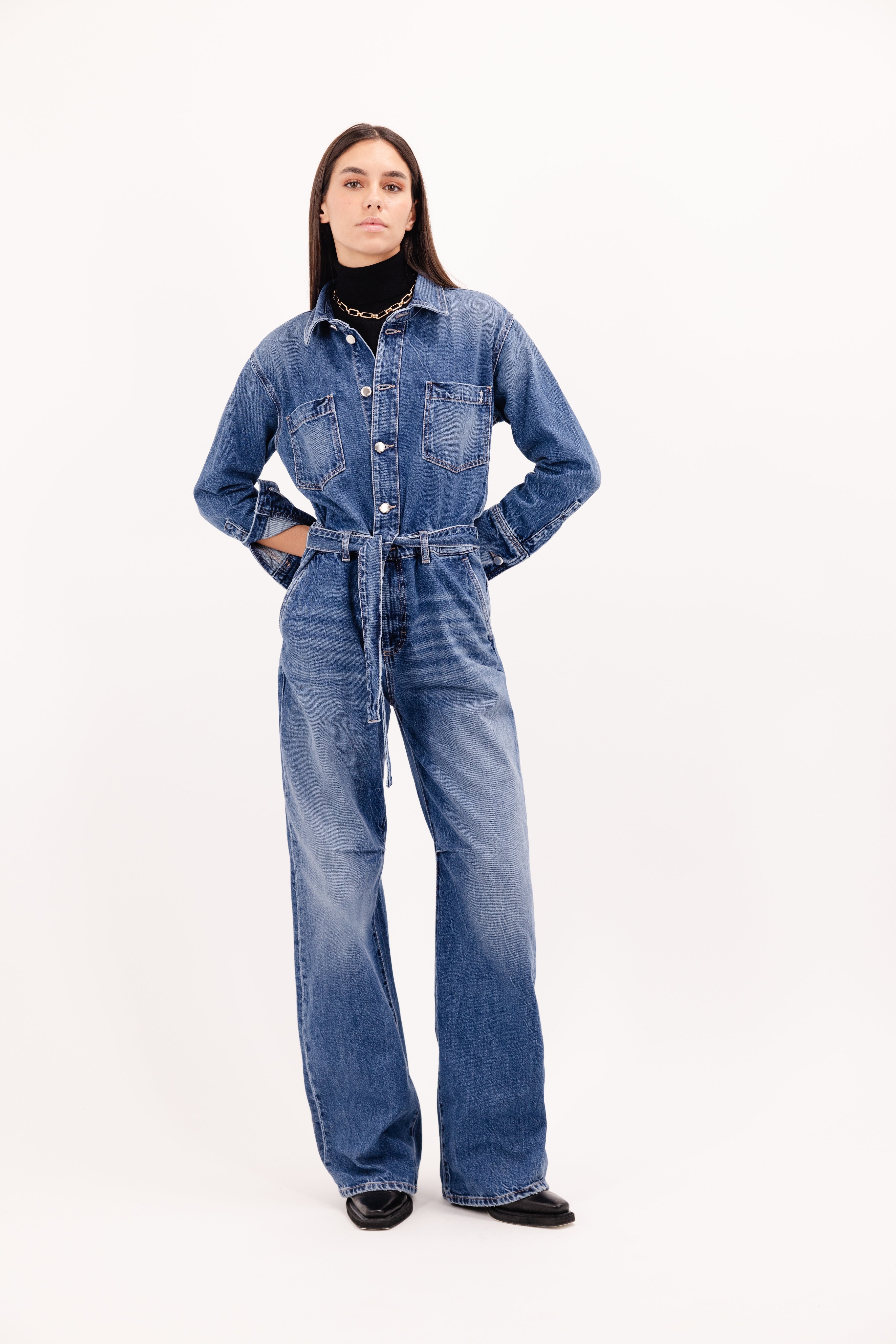 Indie women’s jumpsuit in eclipse wash, 100% rigid cotton denim, worn by a model with a black pullover.
