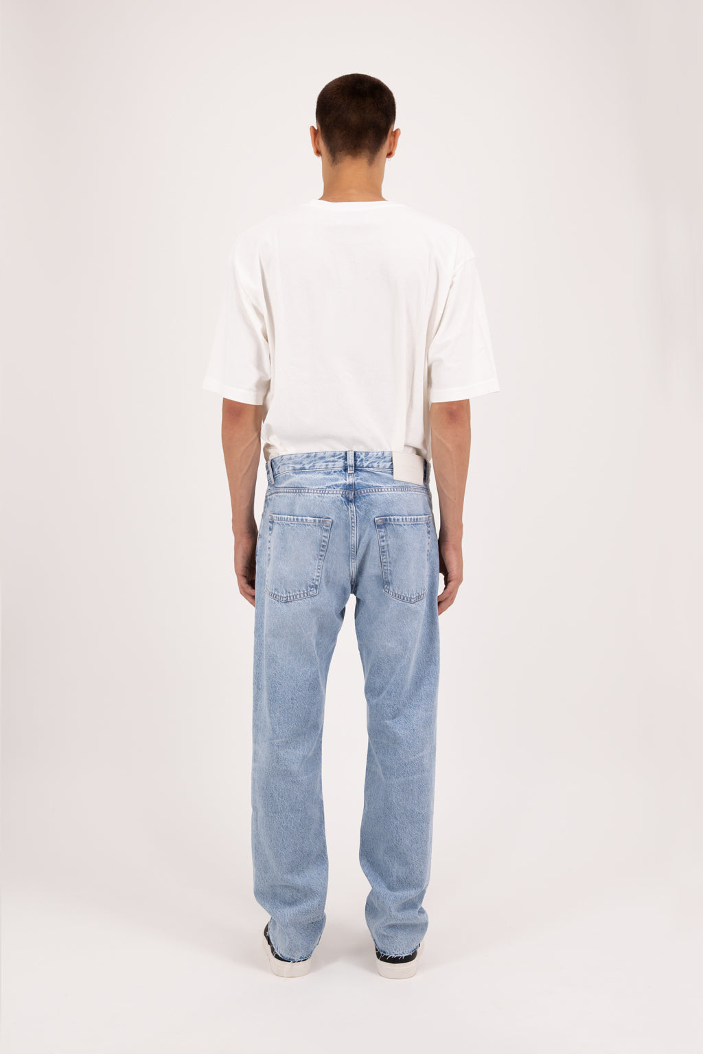 Back view of Kanye Eco men’s baggy jeans in arid blue wash, 100% organic rigid cotton denim, worn by a model with a white t-shirt.