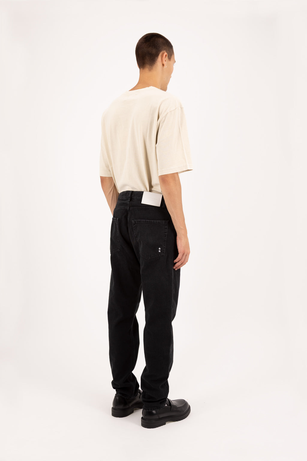 Back view of Kanye men’s baggy jeans in graphite black wash, 100% rigid soft cotton denim, worn by a model with a desert t-shirt.