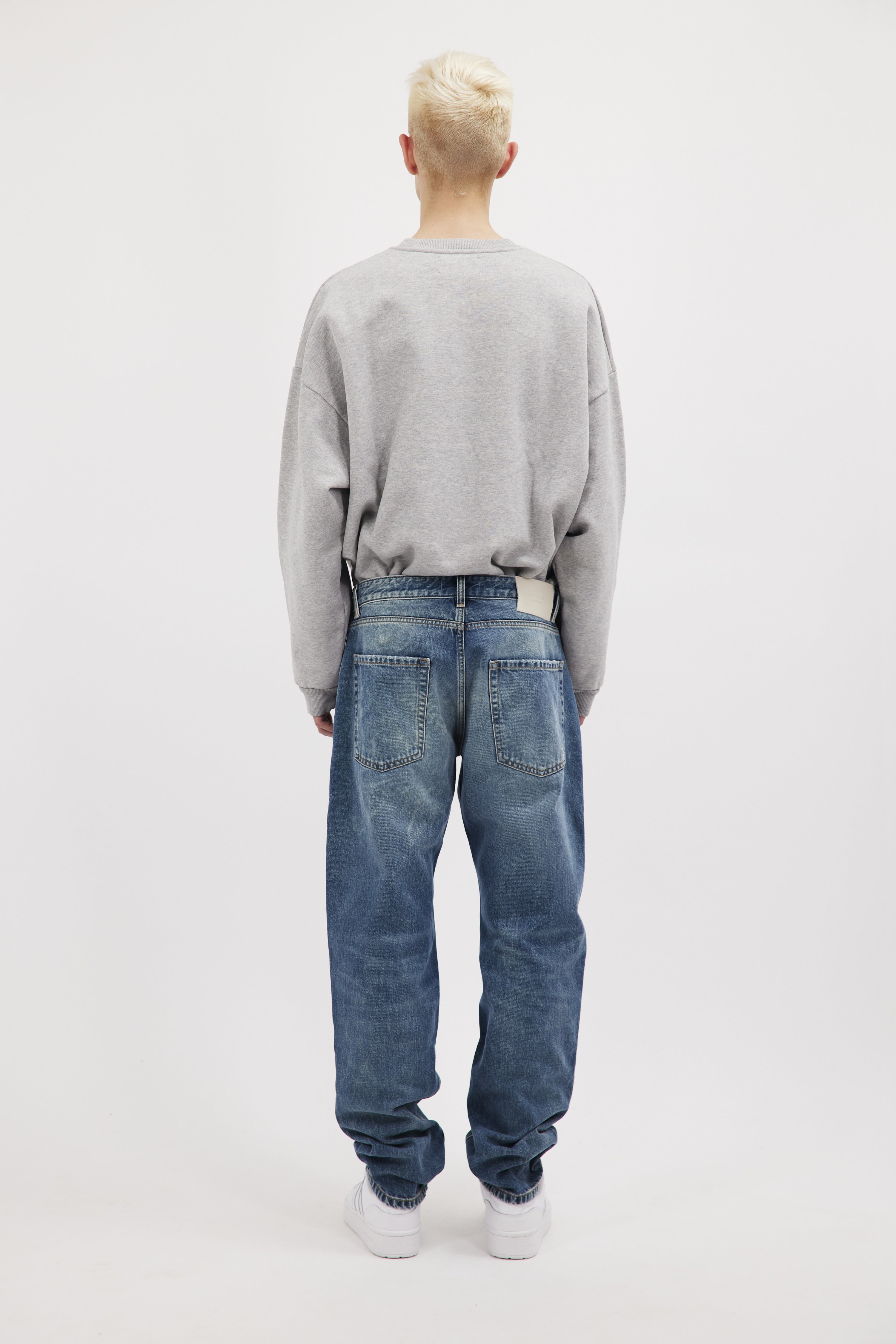 Back view of Kanye men’s loose jeans in aged stone wash, 100% rigid cotton denim, worn by a model with a gray sweatshirt.