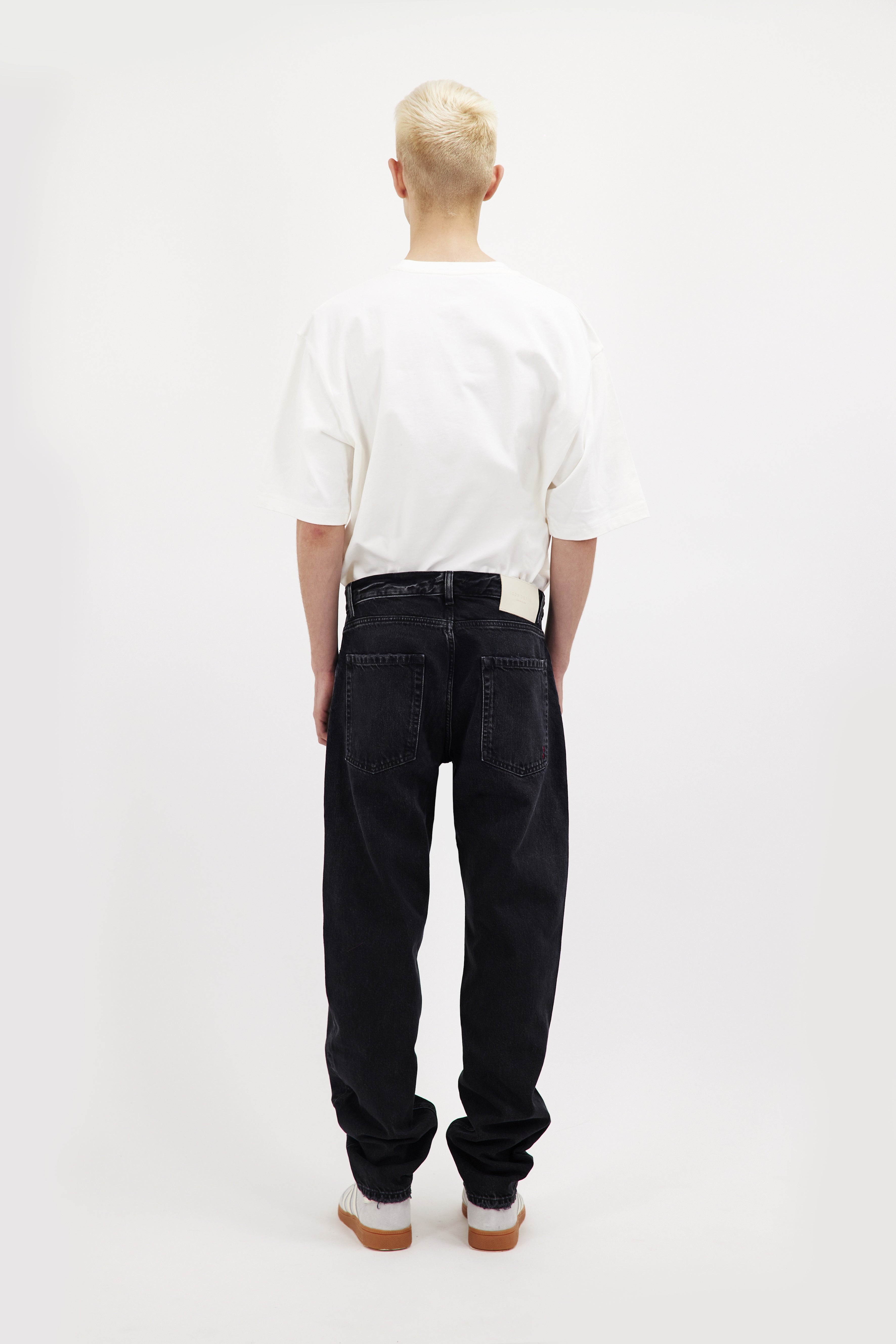 Back view of Kanye men’s loose jeans in graphite black wash, 100% rigid cotton denim, worn by a model with a white t-shirt.