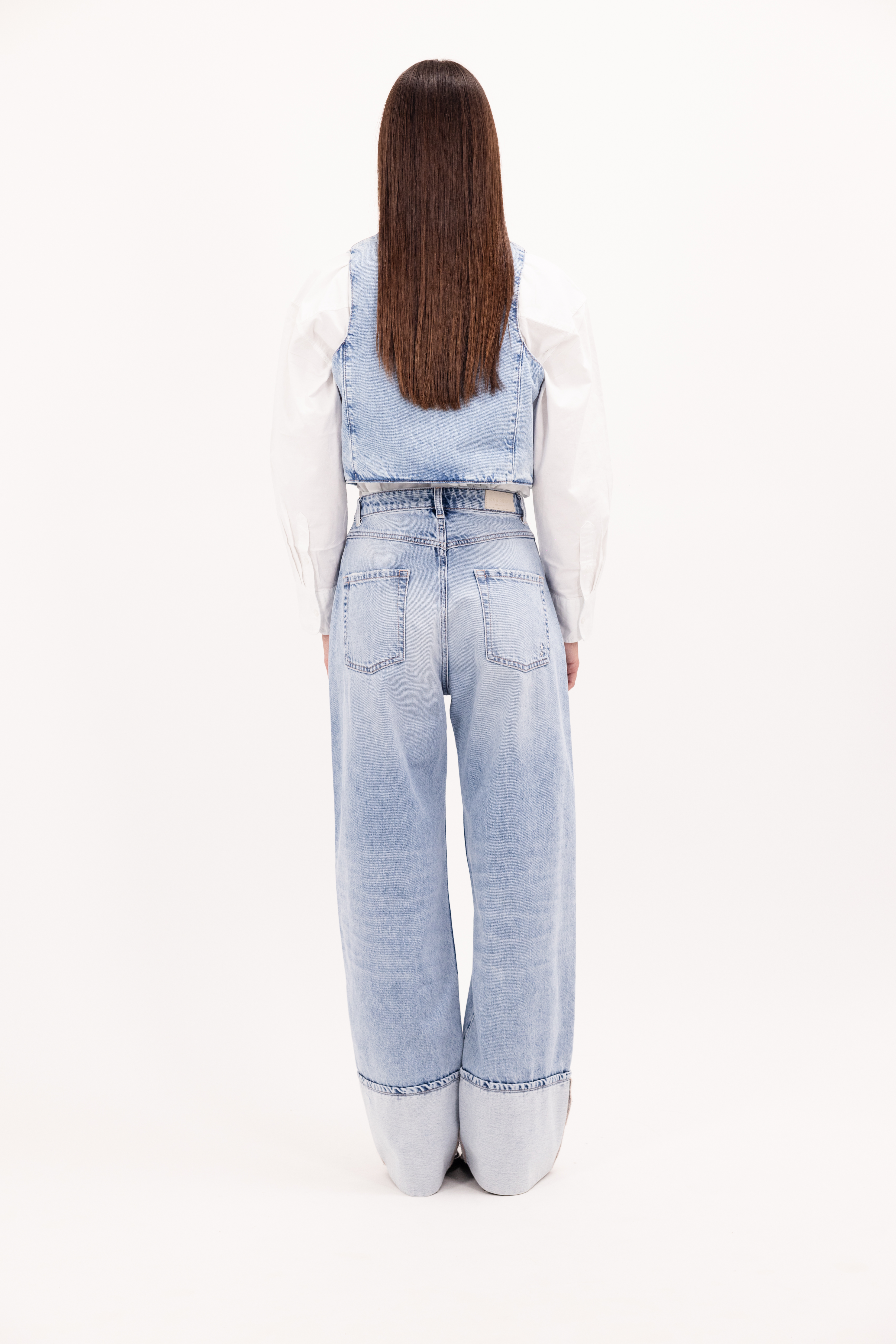 Back view of Kiki women’s wide leg jeans in sky blue wash, 100% rigid cotton denim, worn by a model with a shirt and denim vest.