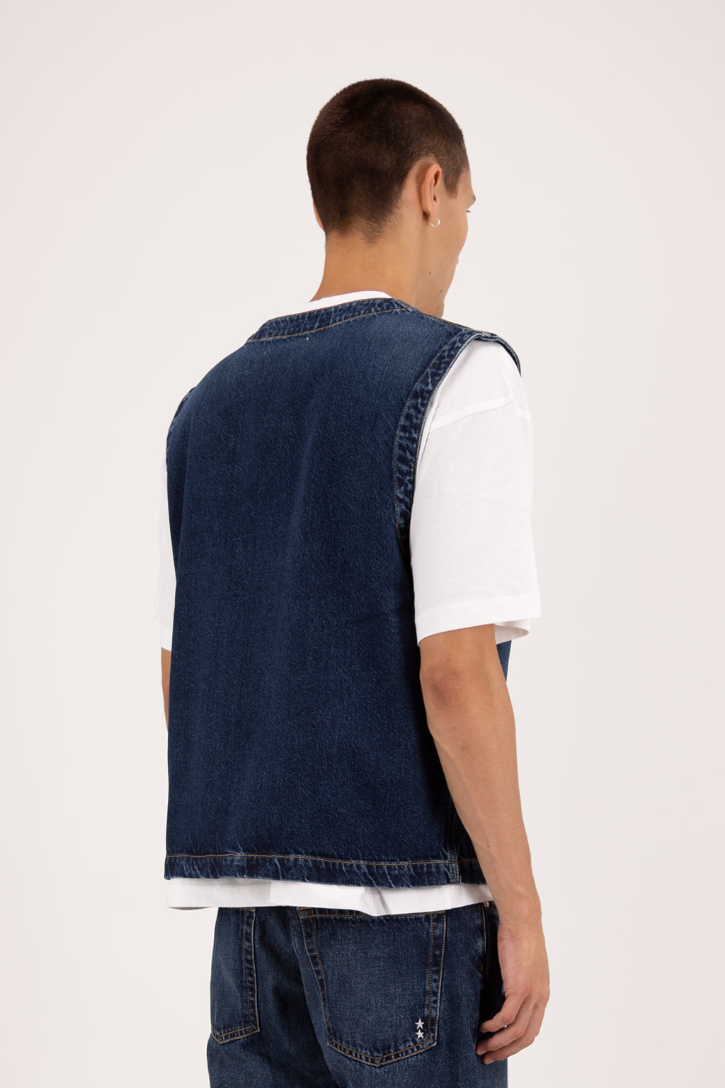 Back view of Kit men’s vest in deep blue wash, 100% rigid cotton denim, worn by a model with a white t-shirt.