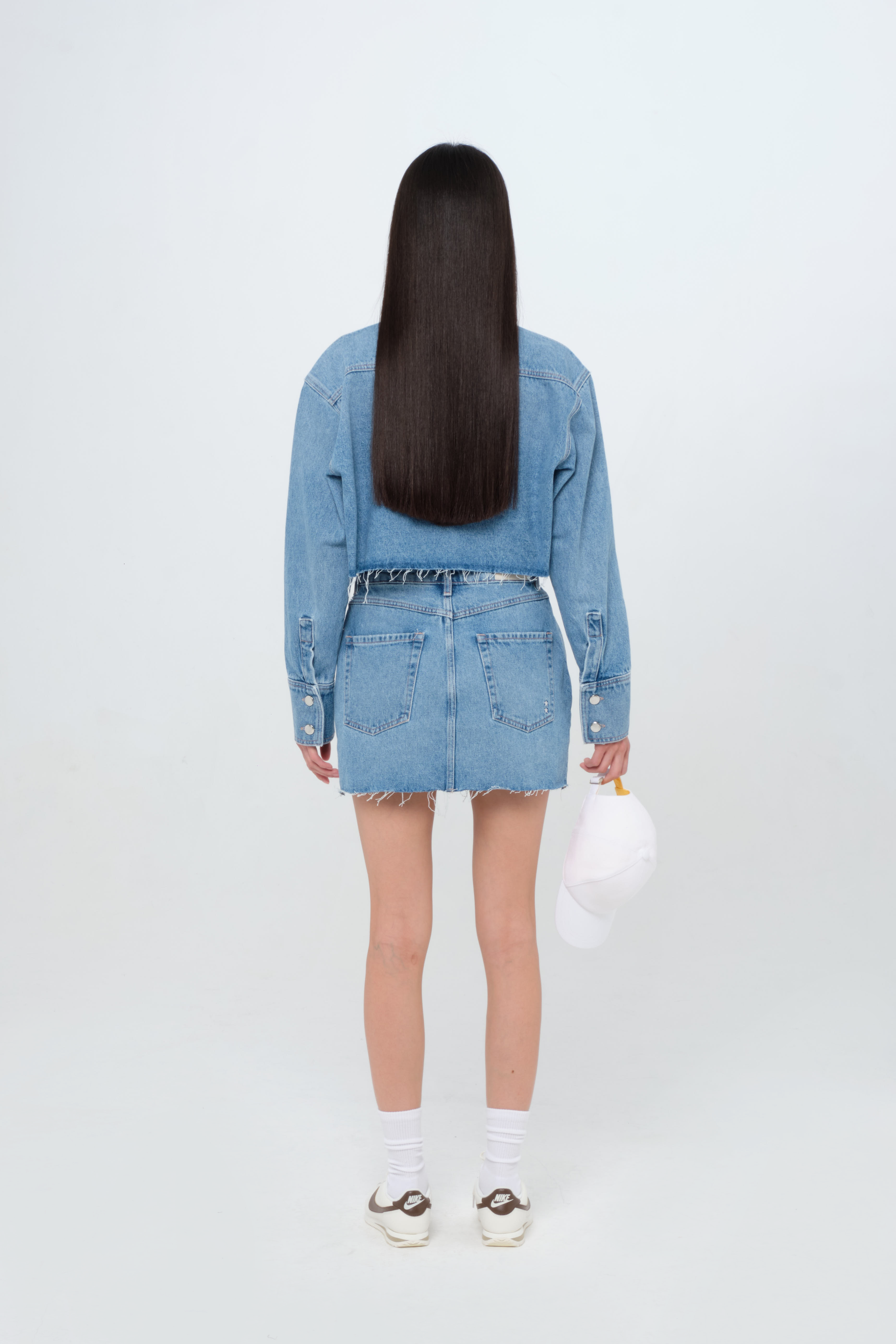 Back view of Nelly Eco women’s short skirt in medium wash, 100% rigid organic cotton denim, worn by a model with a crop denim shirt.