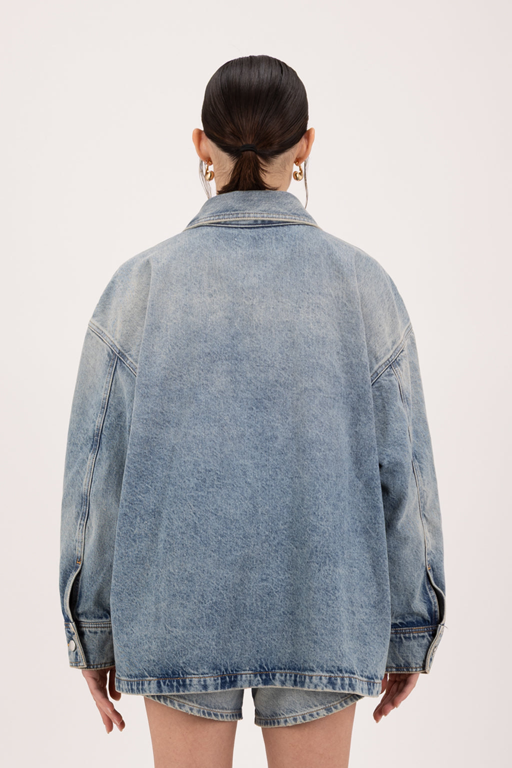 Back view of Nika women’s utility jacket in smoke blue wash, 100% rigid cotton denim, worn by a model with shorts.