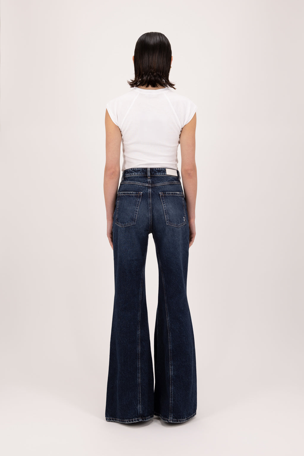 Back view of Stella women’s flared leg jeans in deep blue wash, 100% rigid cotton denim, worn by a model with a white top.
