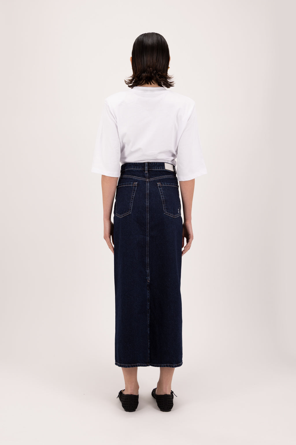 Back view of Suki women’s straight skirt in deep blue wash, 100% rigid cotton denim, worn by a model with a white t-shirt.