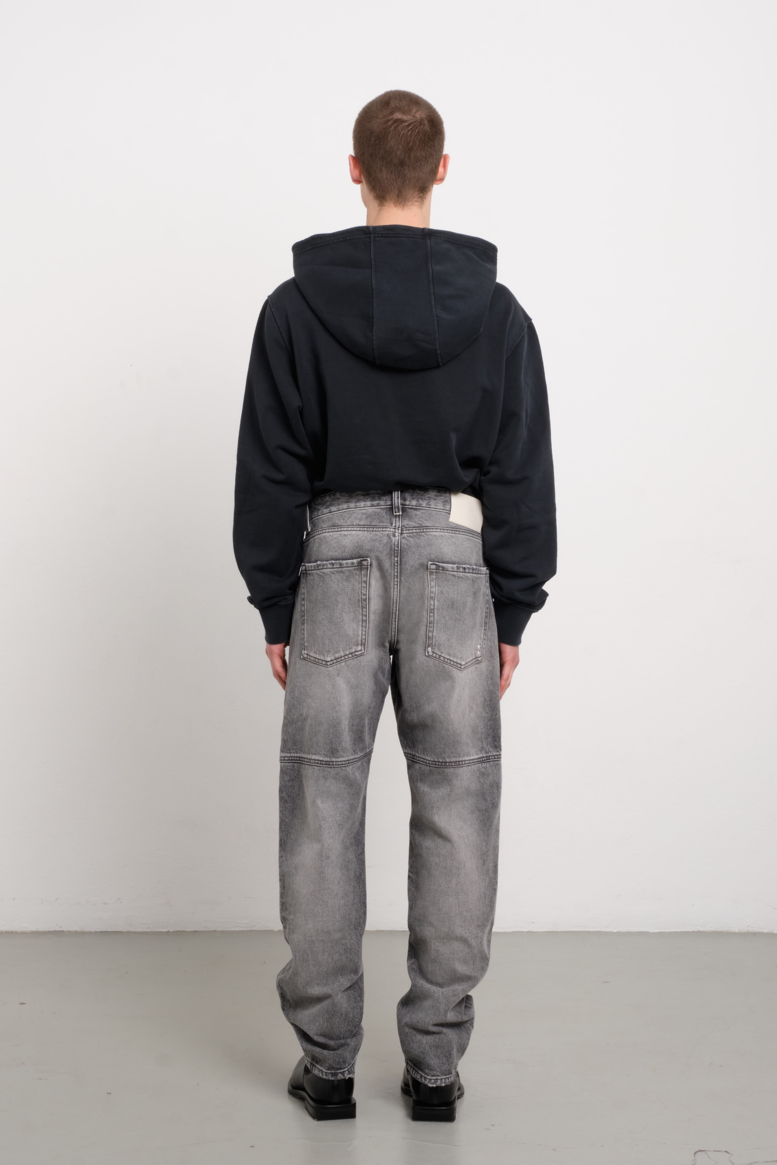Back view of Tavi men’s loose jeans in dark gray wash, 100% rigid cotton denim, worn by a model with a black hoodie.