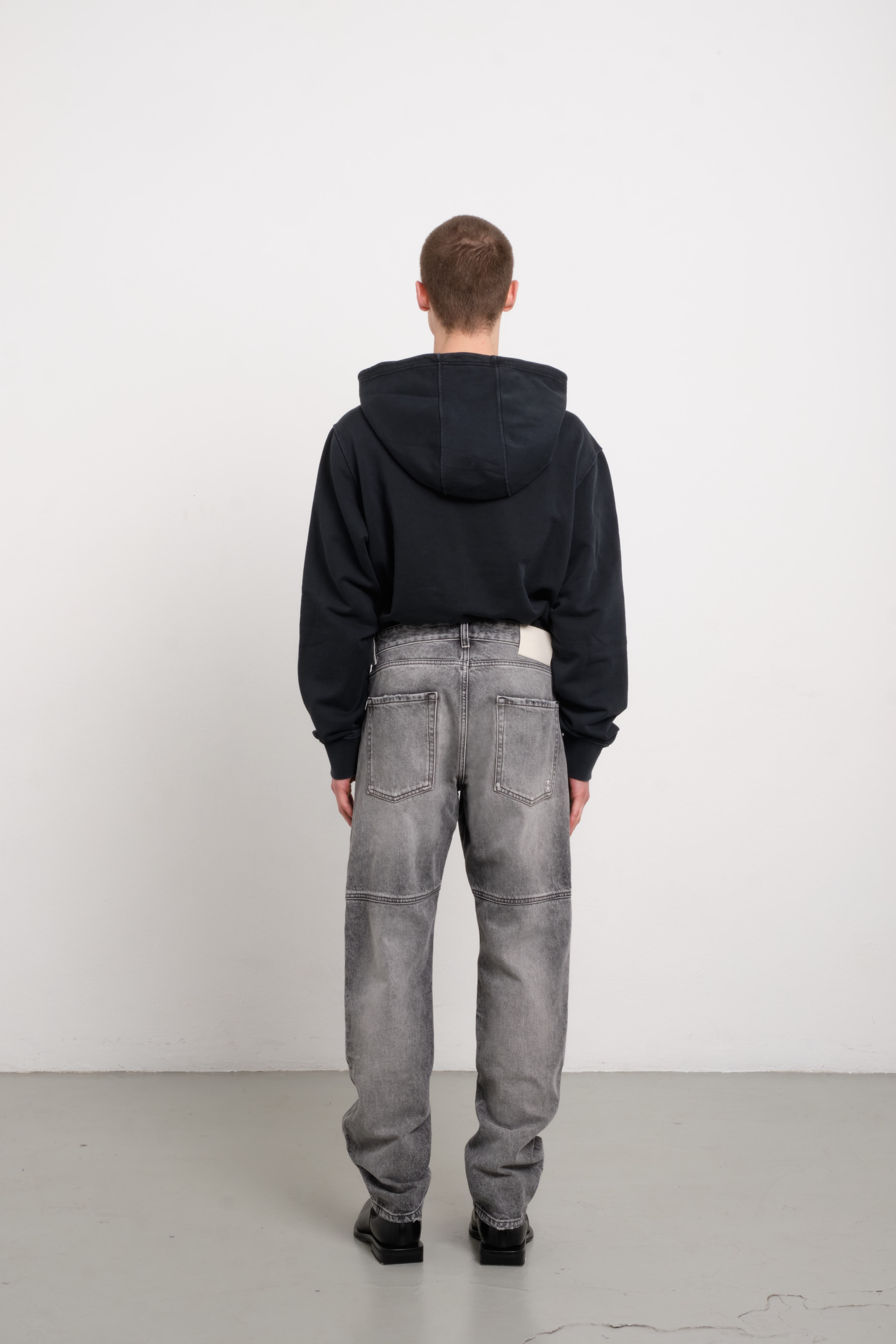 Back view of Tavi men’s loose jeans in dark gray wash, 100% rigid cotton denim, worn by a model with a black hoodie.