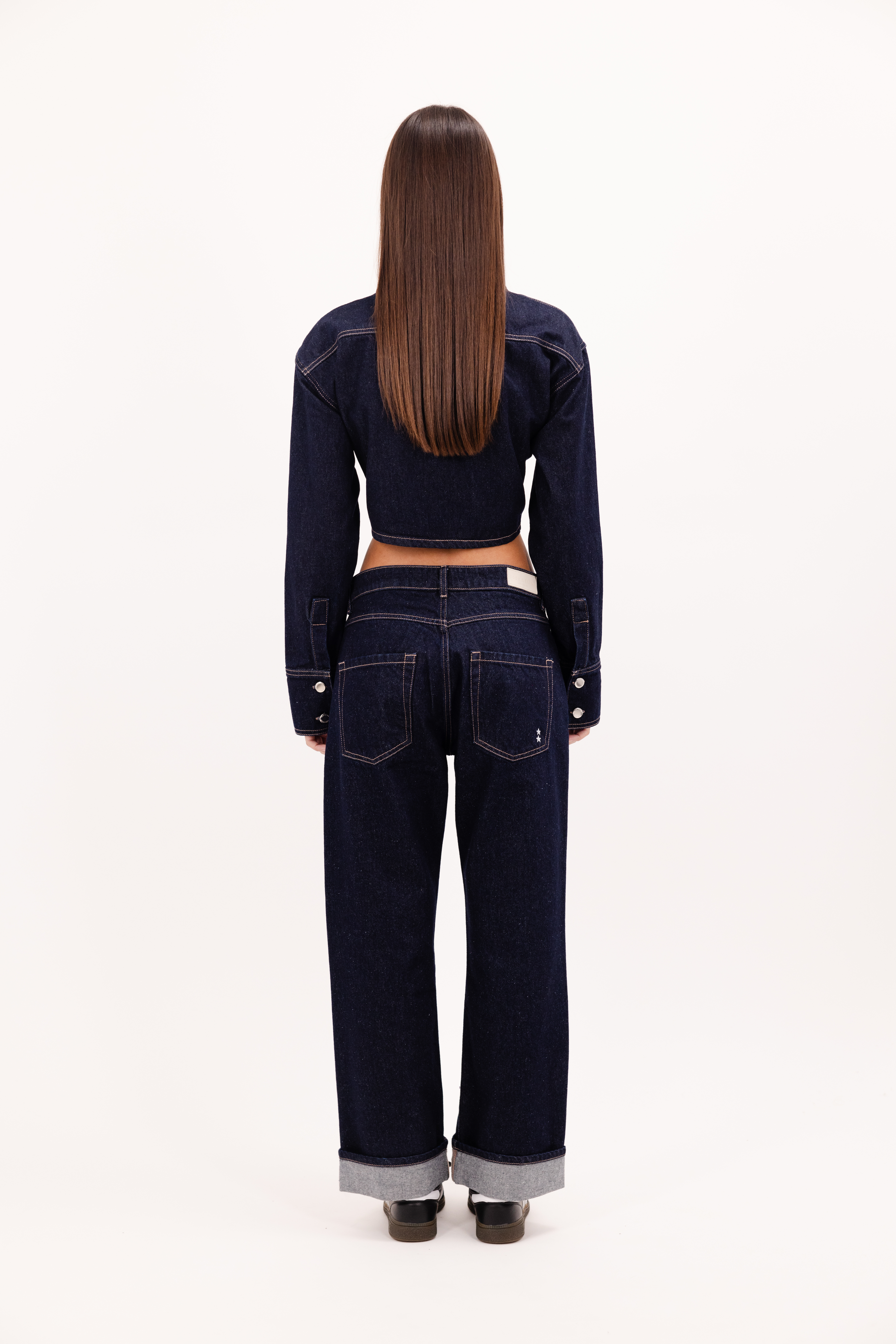 Back view of tiny women’s flared leg jeans in indigo raw, 100% cotton denim, worn by a model with a denim crop shirt.